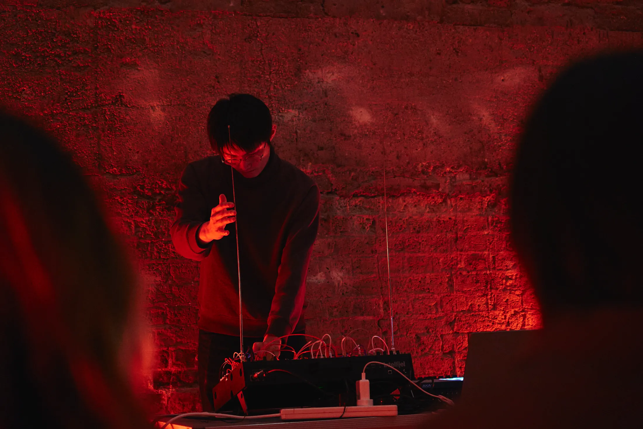 Zhao Jiajing performing live electronics in red light against a stone wall, one hand raised beside a slender antenna above a modular synthesiser